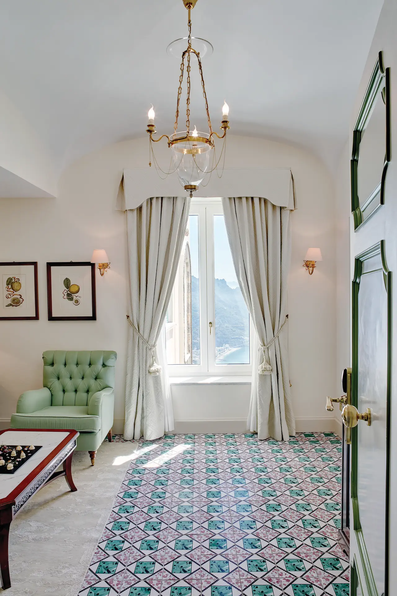 The sitting room of a Deluxe Sea View Terrace Suite: the green majolica floor, a buttoned armchair and the curtained window.
