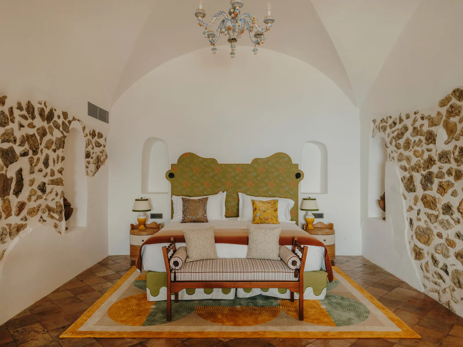 A Divina Suite bedroom: the bed with its green headboard between 2 bare stone walls, under a glass chandelier.
