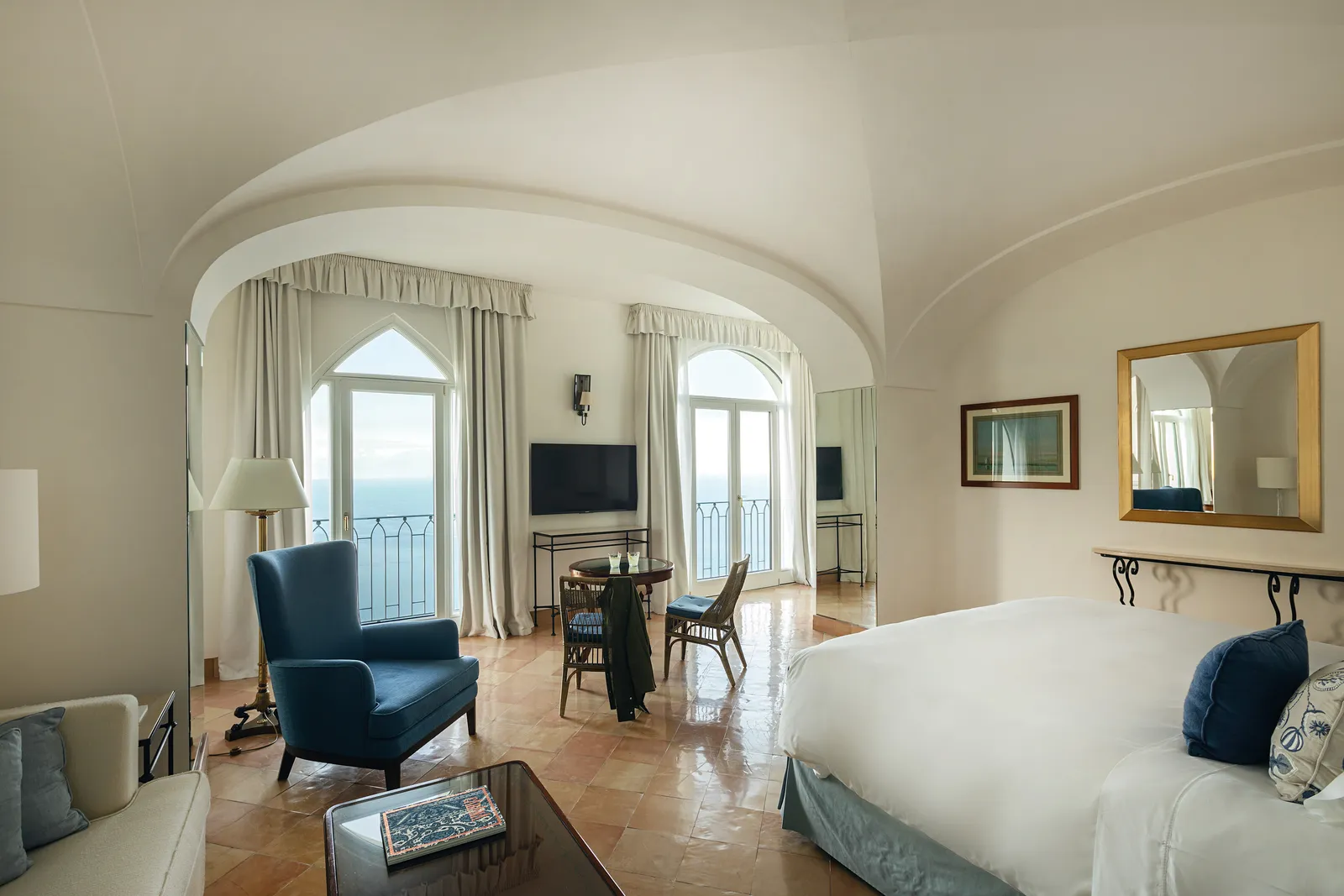 A Deluxe Sea View Junior Suite with Balcony: the bed, a blue armchair and the arched windows open to the sea.