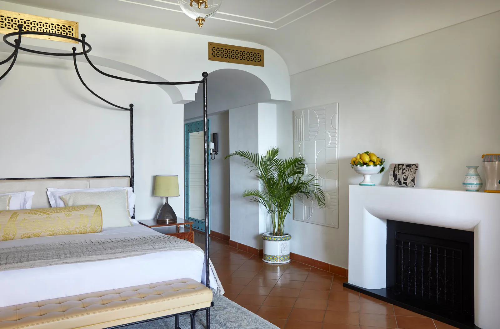 A Sea View Suite with Garden: the iron four-poster bed, the white fireplace and a potted palm.