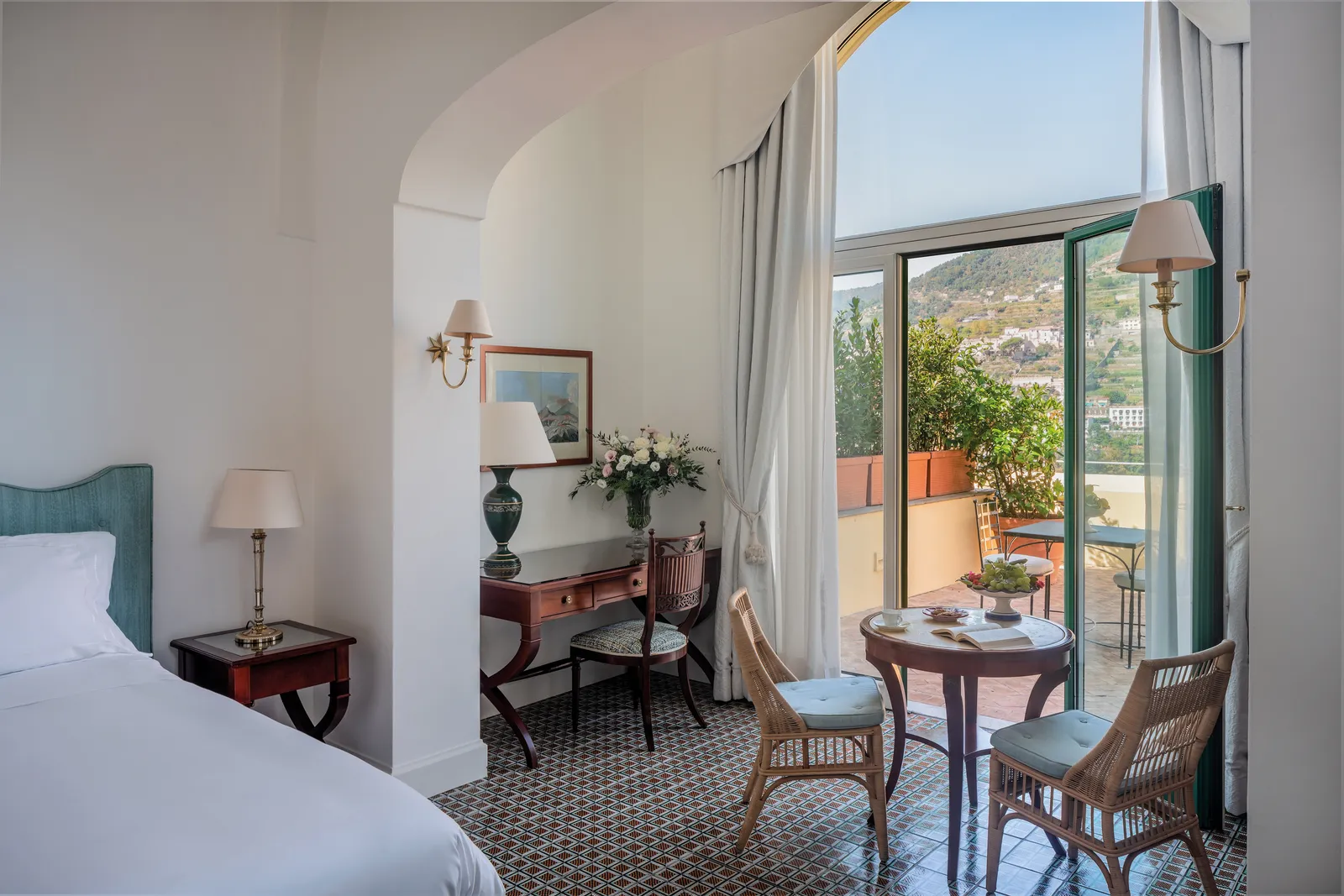 A Superior Room with Garden: the bed, a small table with wicker chairs and the French window open onto the terrace and the village.