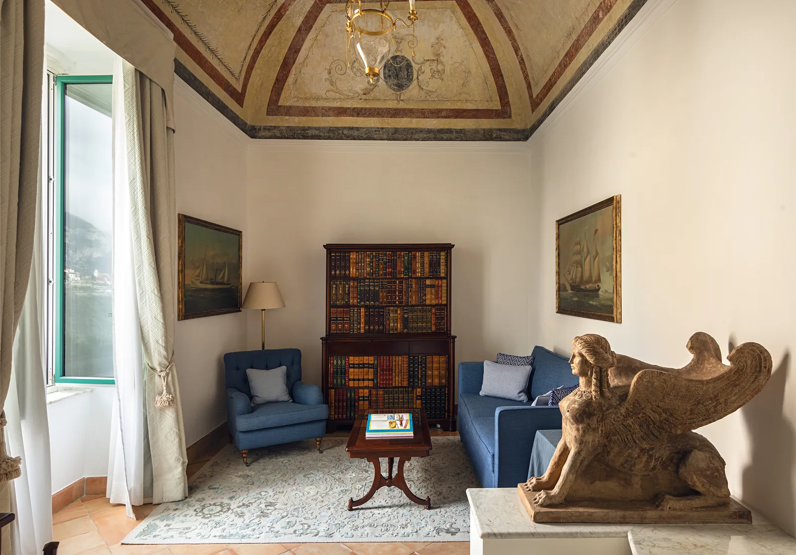 The sitting room of a Superior Suite: a winged terracotta sphinx, the blue sofa, a bookcase and the frescoed ceiling.
