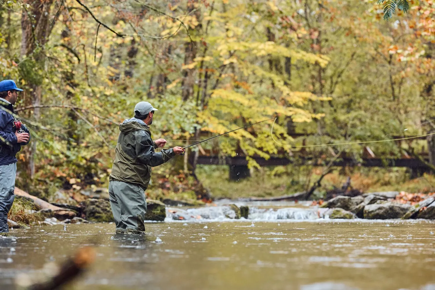 2 fly fishers standing in the shallows of a creek, among boulders and yellow autumn trees.