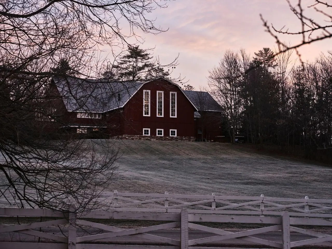 A red barn behind a white fence at dawn, the field covered in frost and bare trees against a pink sky.