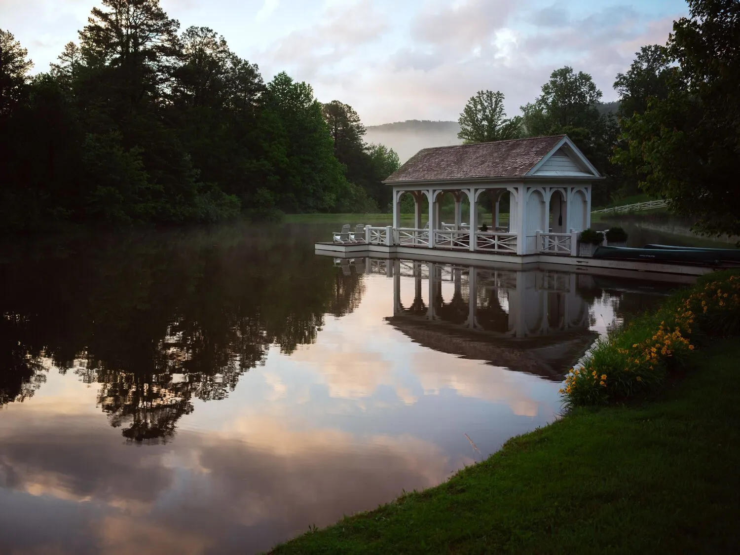 The white boathouse pavilion on the pond at dusk, pink clouds reflected in the still water and woods all around.