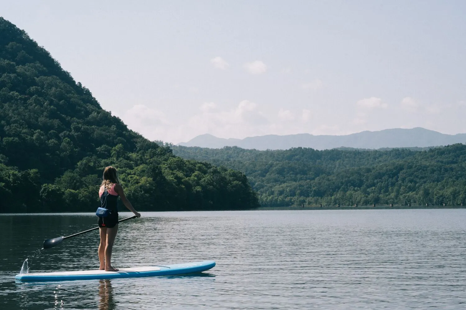 A woman standing on a paddleboard on a calm lake, between wooded slopes with blue mountains in the distance.