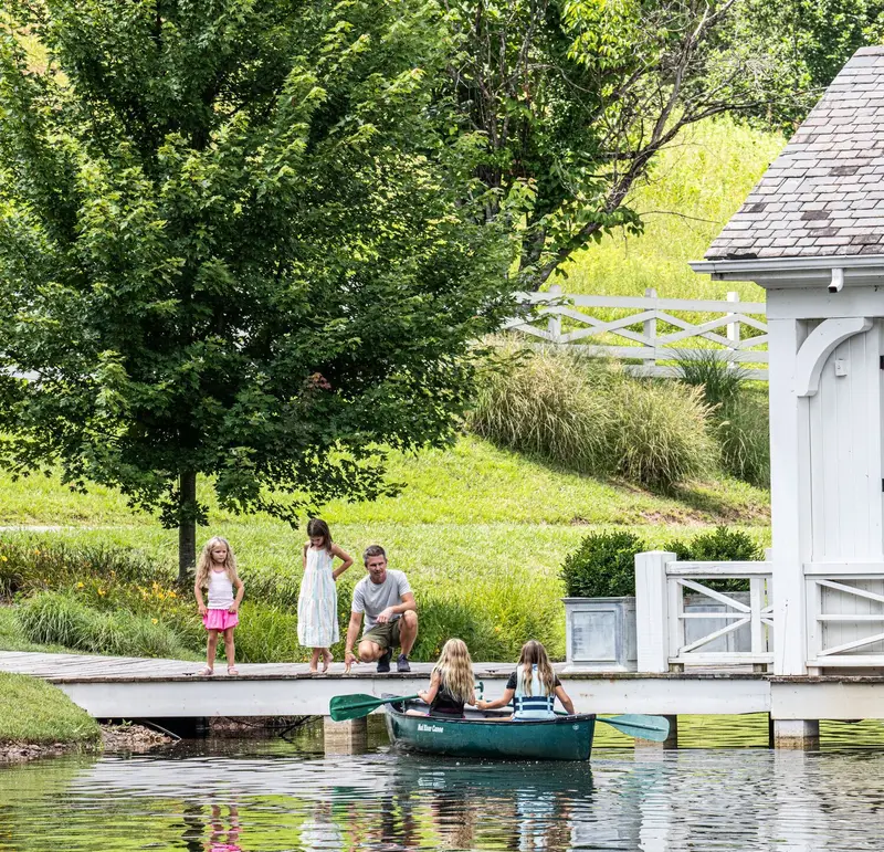 2 girls in a canoe on the pond beside the white boathouse’s jetty, an adult and 2 more girls watching from the bank.