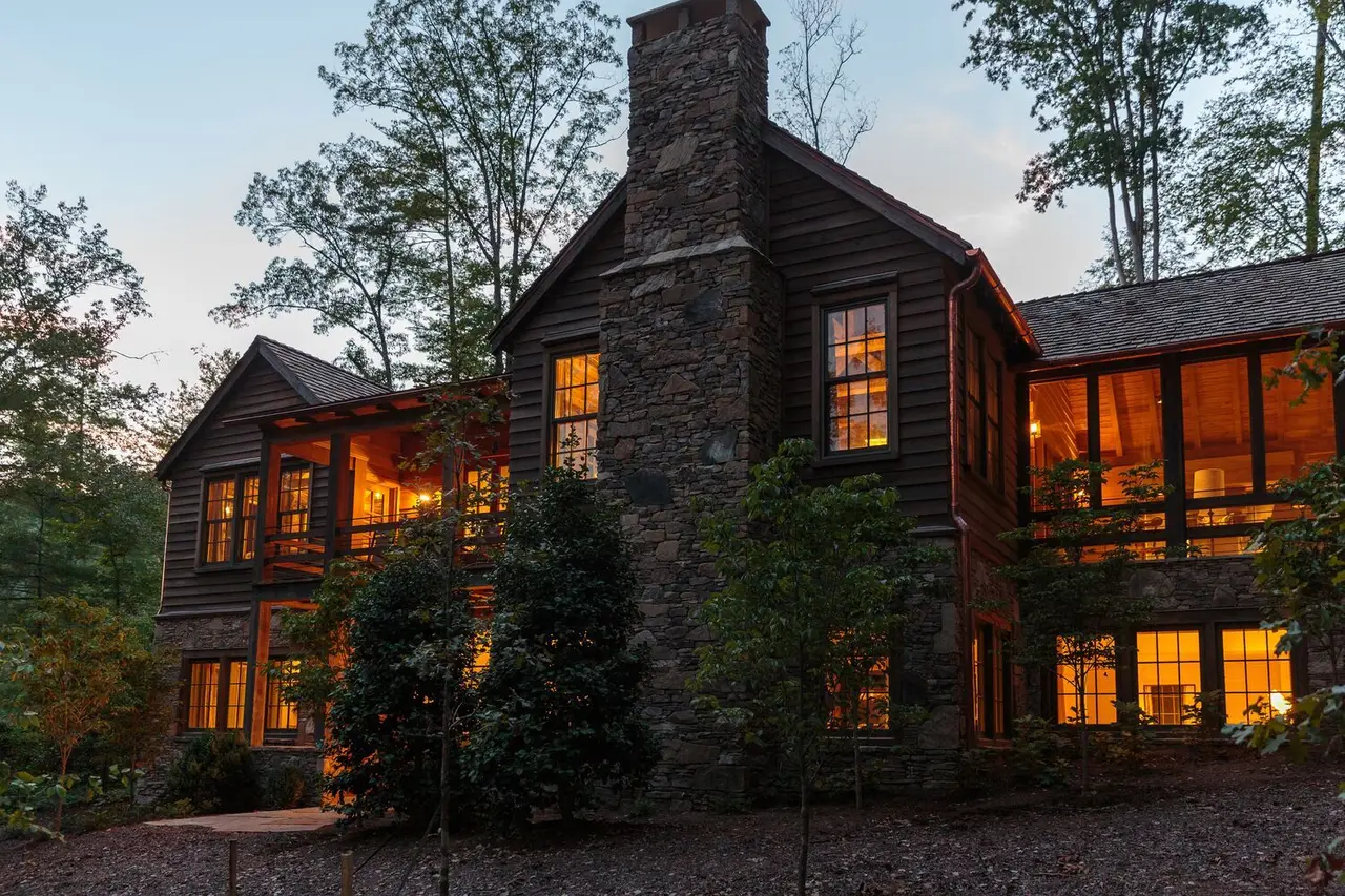 American Rob Mill at dusk: the dark timber house with its stone chimney and lit windows among the trees.