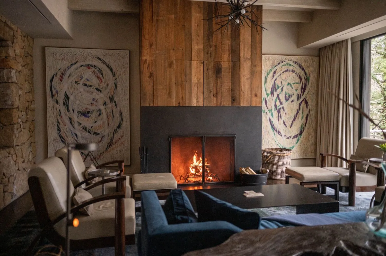 Boulder Cottage’s living room: the fire lit in a wall of rough timber, 2 large abstract canvases and armchairs round the low table.