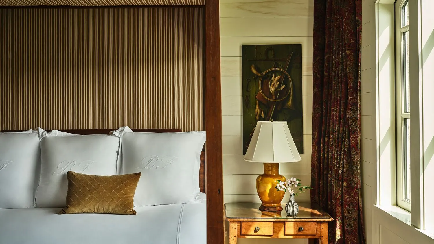 A Carriage House Suite’s bed, with a dark striped headboard, monogrammed pillows and a gilt lamp on the bedside table by the window.