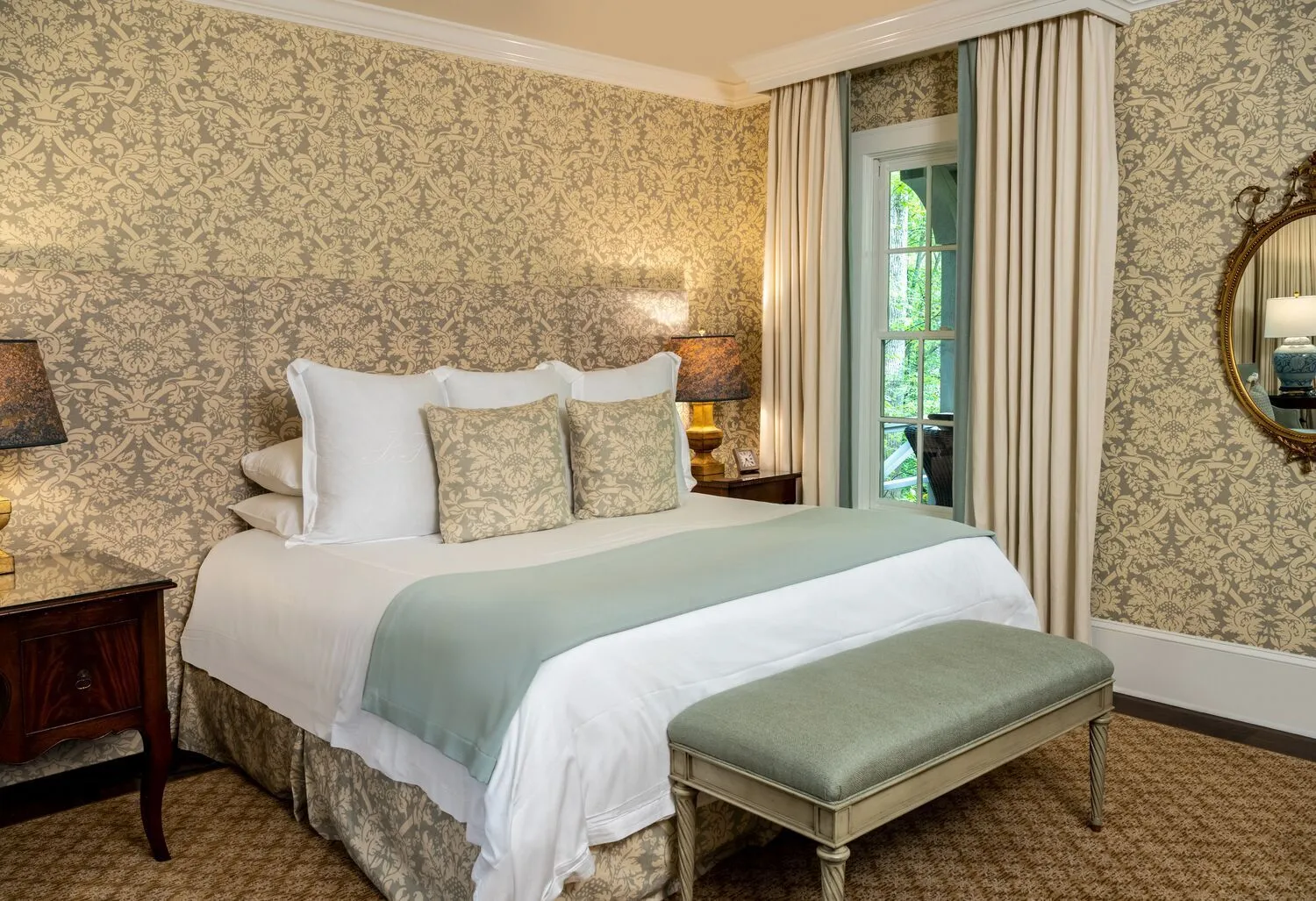 A Holly Glade Cottage Suite’s bedroom: damask wallpaper, a king bed with a sage-green throw and a bench at its foot.