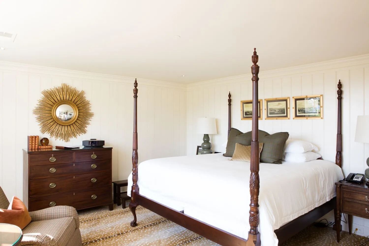 An Estate Room in the Guest House: white panelled walls, a mahogany four-poster, a chest of drawers and a gilt sunburst mirror.