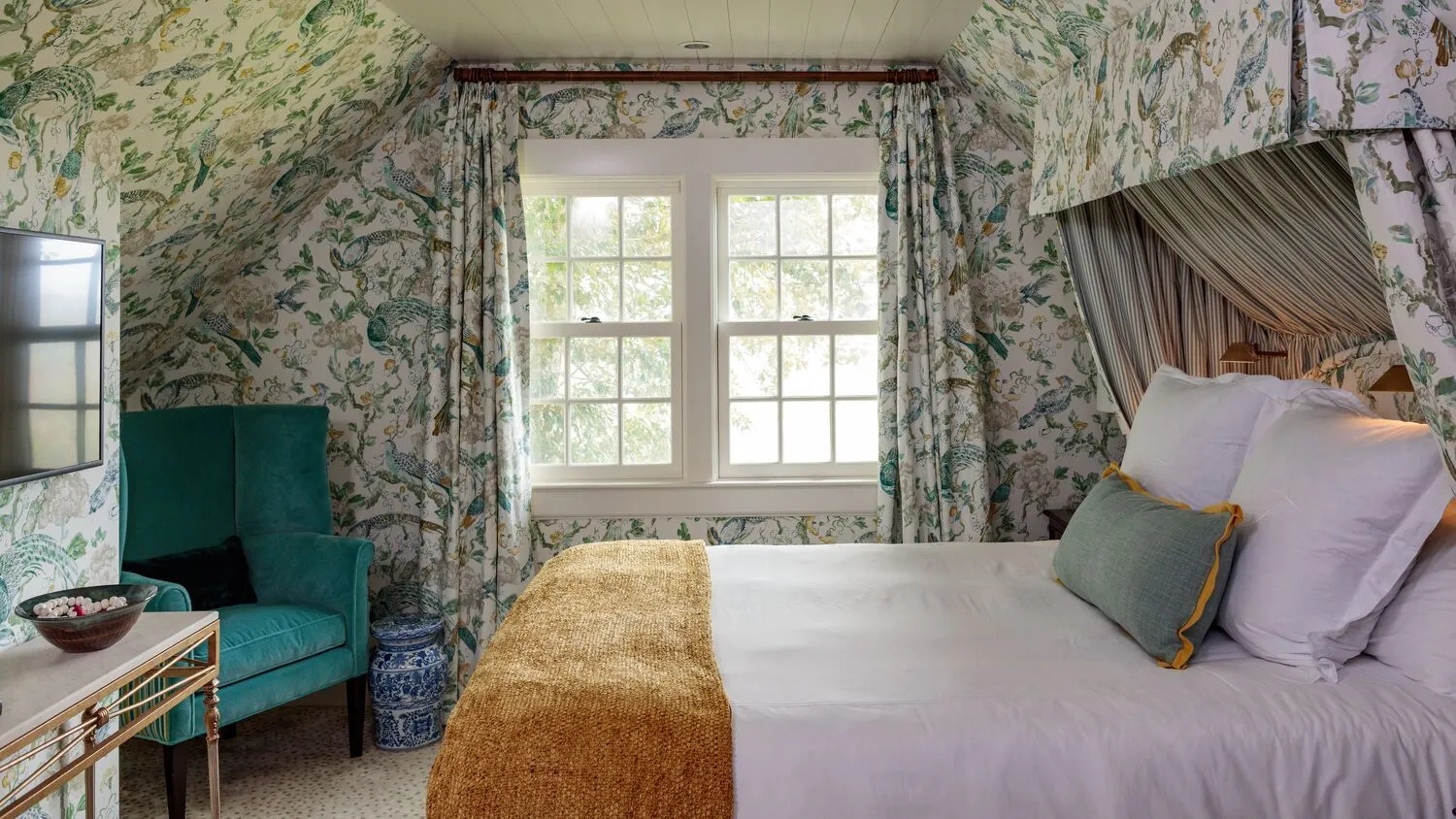 A Historic Room under the eaves of the Main House: green floral wallpaper, a fabric canopy over the bed and a green velvet armchair.