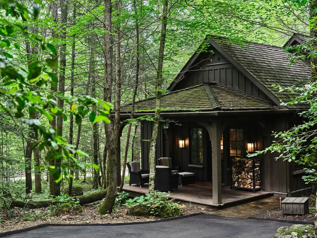A Singing Brook cottage among the trees: the dark timber house with its mossy roof, the porch with armchairs and stacked firewood.