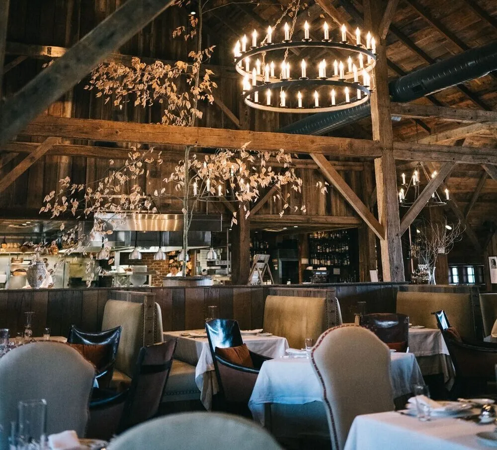 The Barn’s dining room: the old barn’s timbers, a great candle chandelier, flowering branches and white-clothed tables in front of the open kitchen.