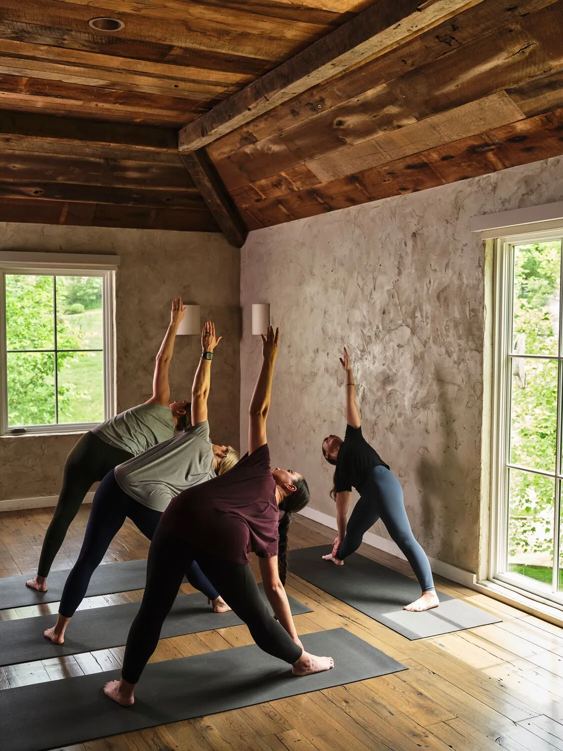 A yoga class at the Wellhouse: 4 people in a pose on dark mats under a timber ceiling, light coming through the windows.