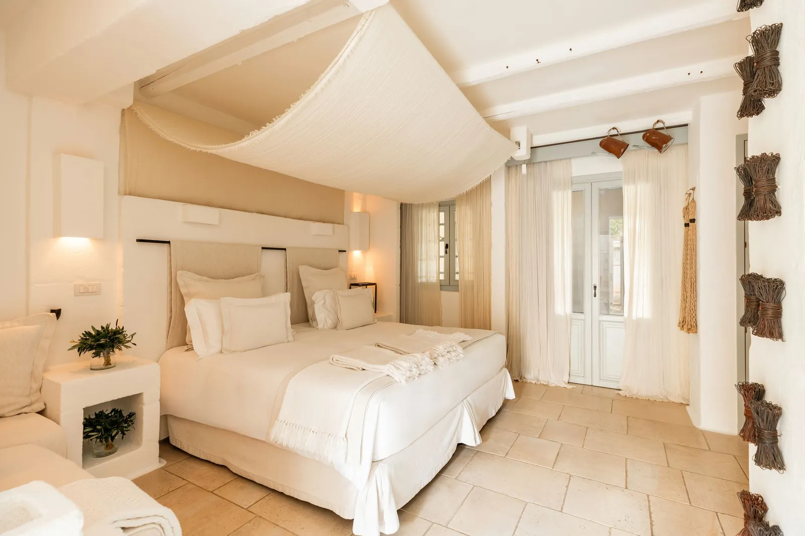 A Borgo Bella room: a large bed under a draped canopy, a built-in bedside table with olive sprigs in a glass vase and a glazed door with sheer curtains.