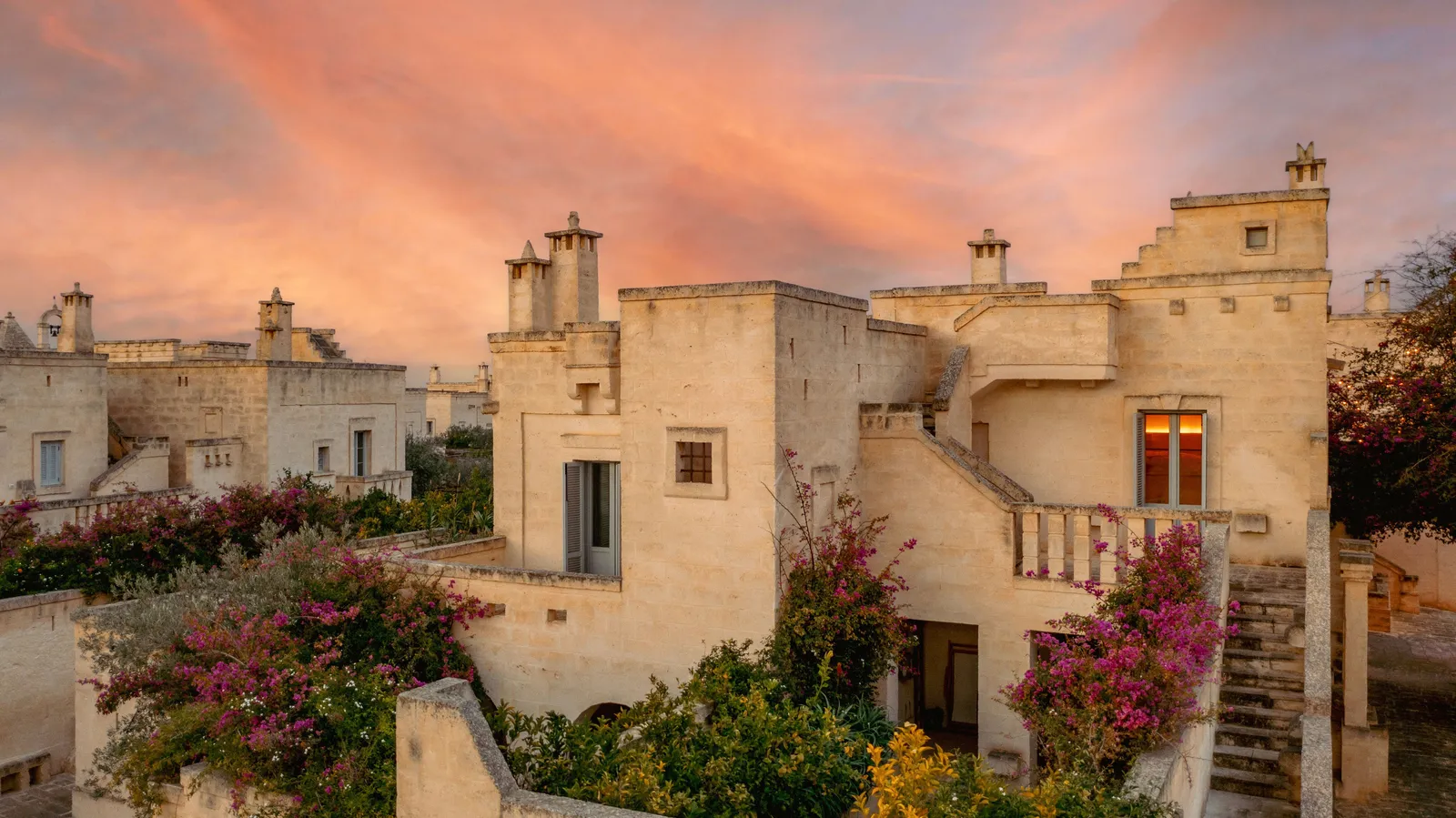 Stone houses in Il Borgo with chimneys, outside stairs and bougainvillea under a pink sky; one window is lit.