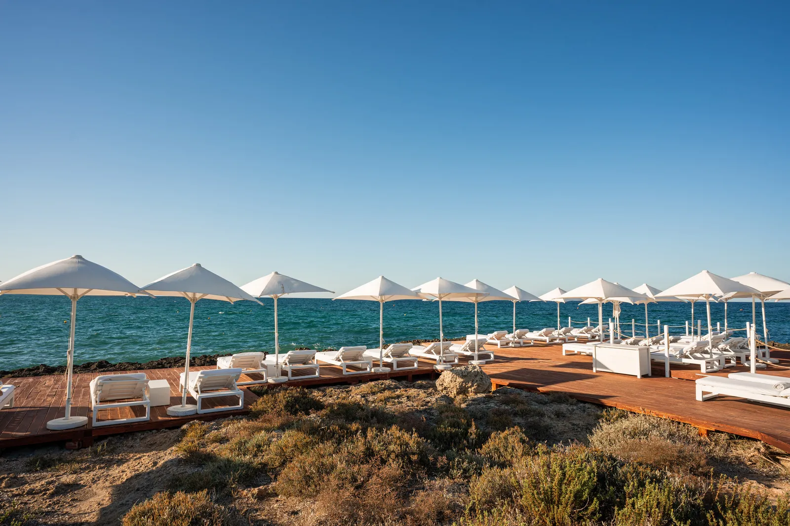 Cala Masciola beach club: a wooden deck with white parasols and loungers on the rocky shore, facing the sea.