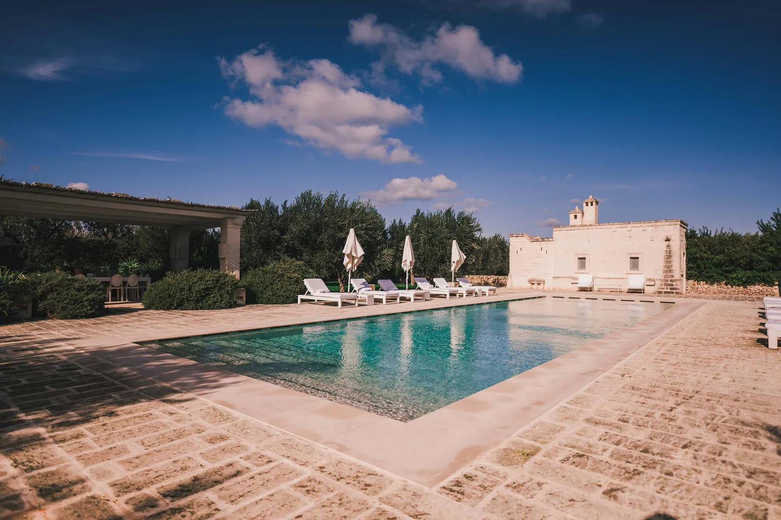 The Casa Padronale pool: a long pool with loungers and parasols, a pergola on the left and a white building at the far end.