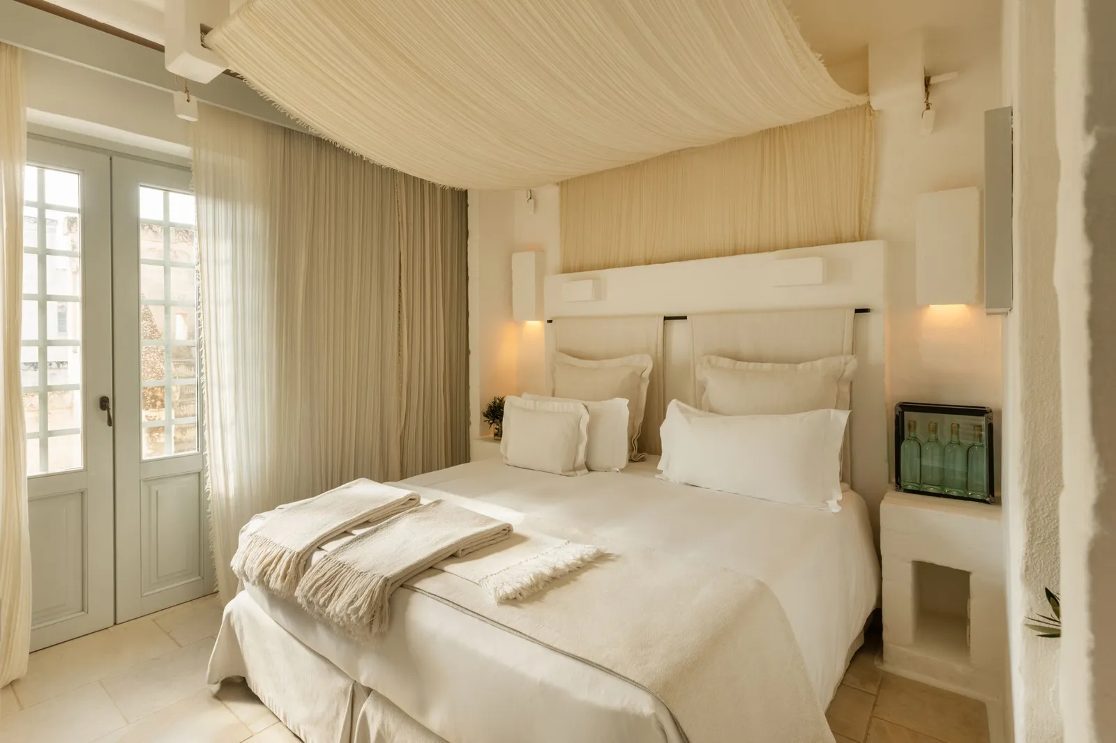 A Casetta Magnifica bedroom: bed under a draped canopy, built-in bedside tables with glass lamps and glazed French windows with long curtains.