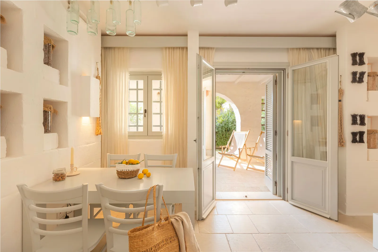 The living room of a Casetta Splendida: white table and chairs, a basket of lemons, stone niches and glass doors open onto a patio with deckchairs.