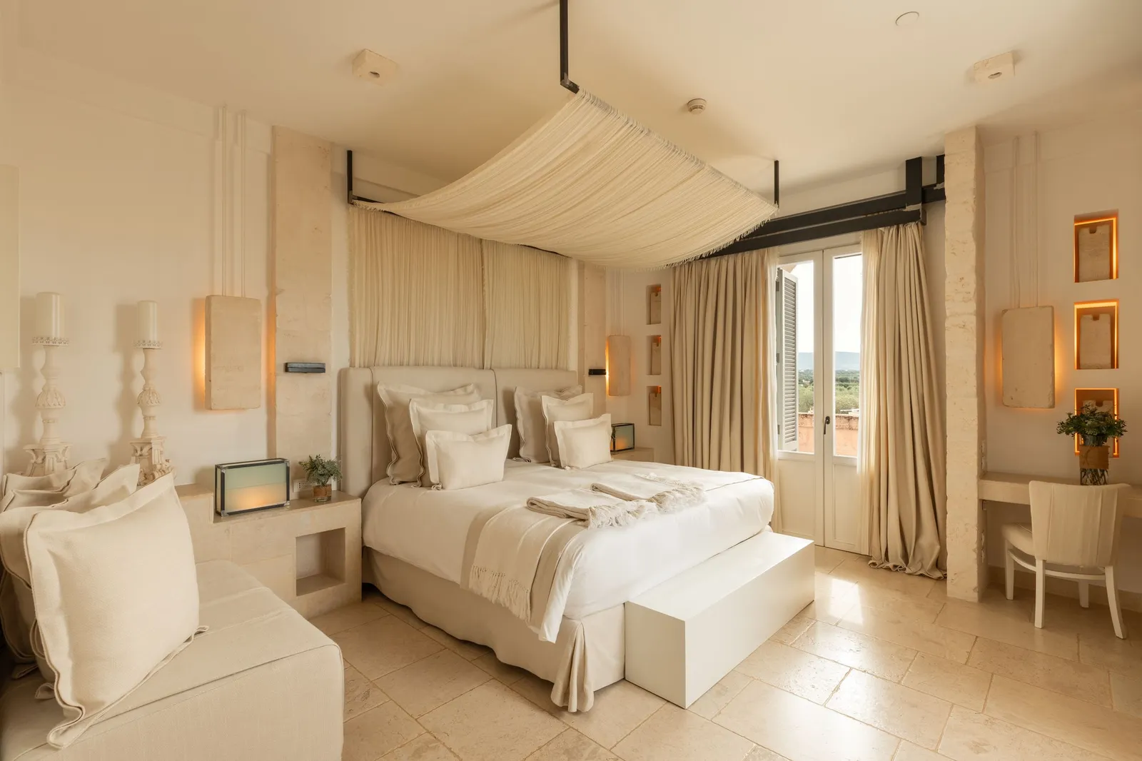 A La Corte Magnifica room: bed under a draped canopy, a sofa, a desk and French windows with long curtains.