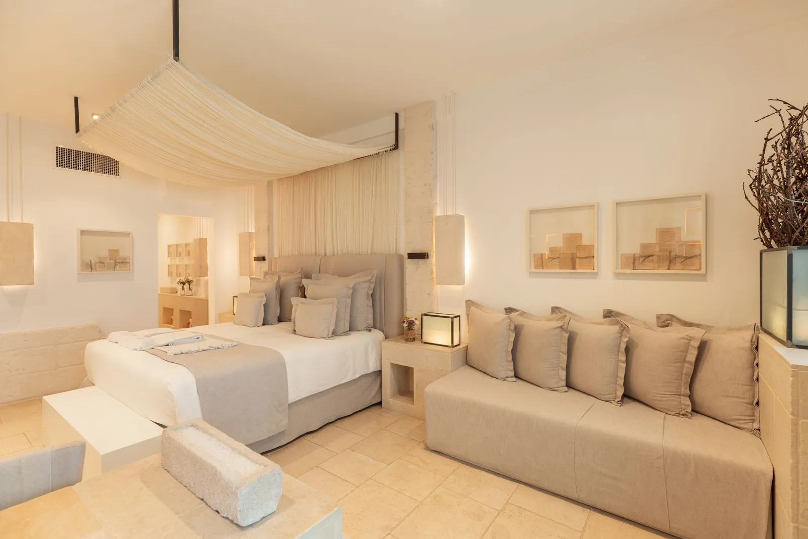 A La Corte Splendida room: bed under a draped canopy, a long sofa with cushions, framed prints and a vase of dry branches.
