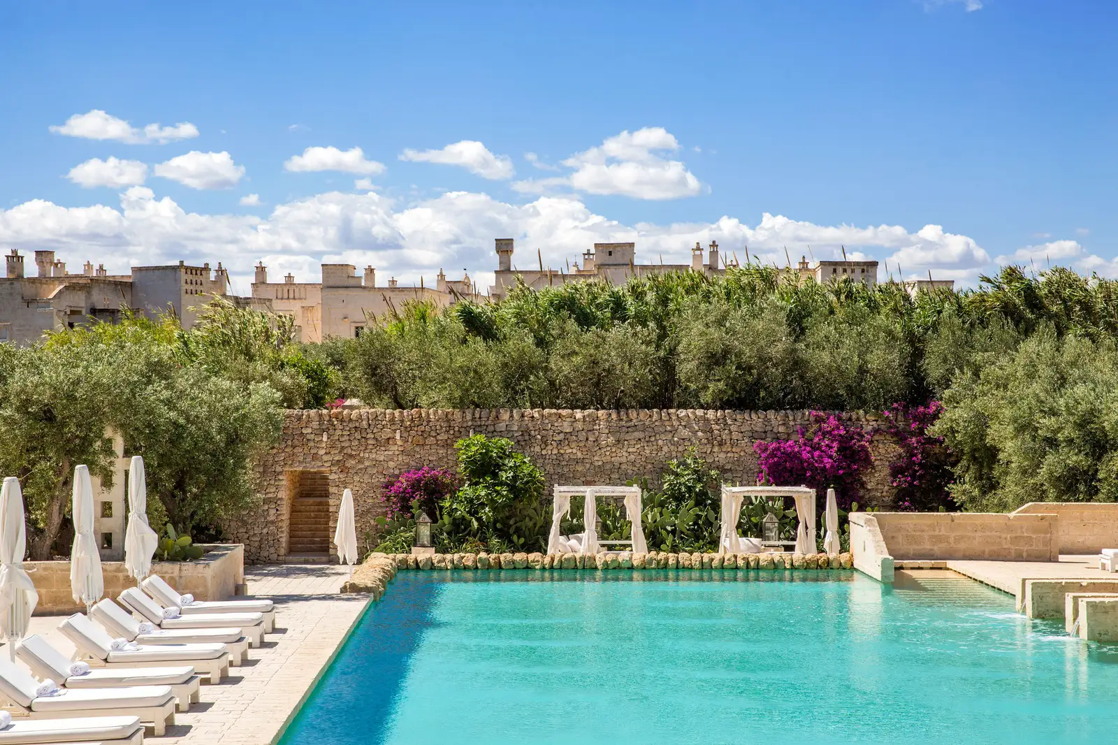 One of La Corte’s pools by day: white loungers and parasols, 2 canopied daybeds, a stone wall with bougainvillea and stone buildings beyond the trees.