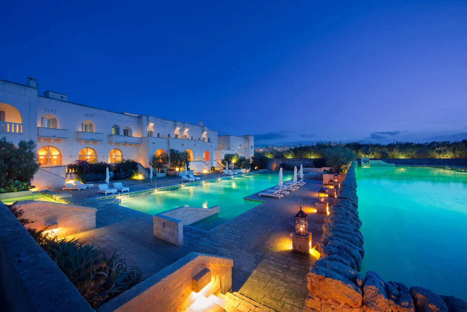 La Corte’s pools at twilight: the lit arches of the main building, loungers by the water and lanterns along the stone wall between 2 pools.