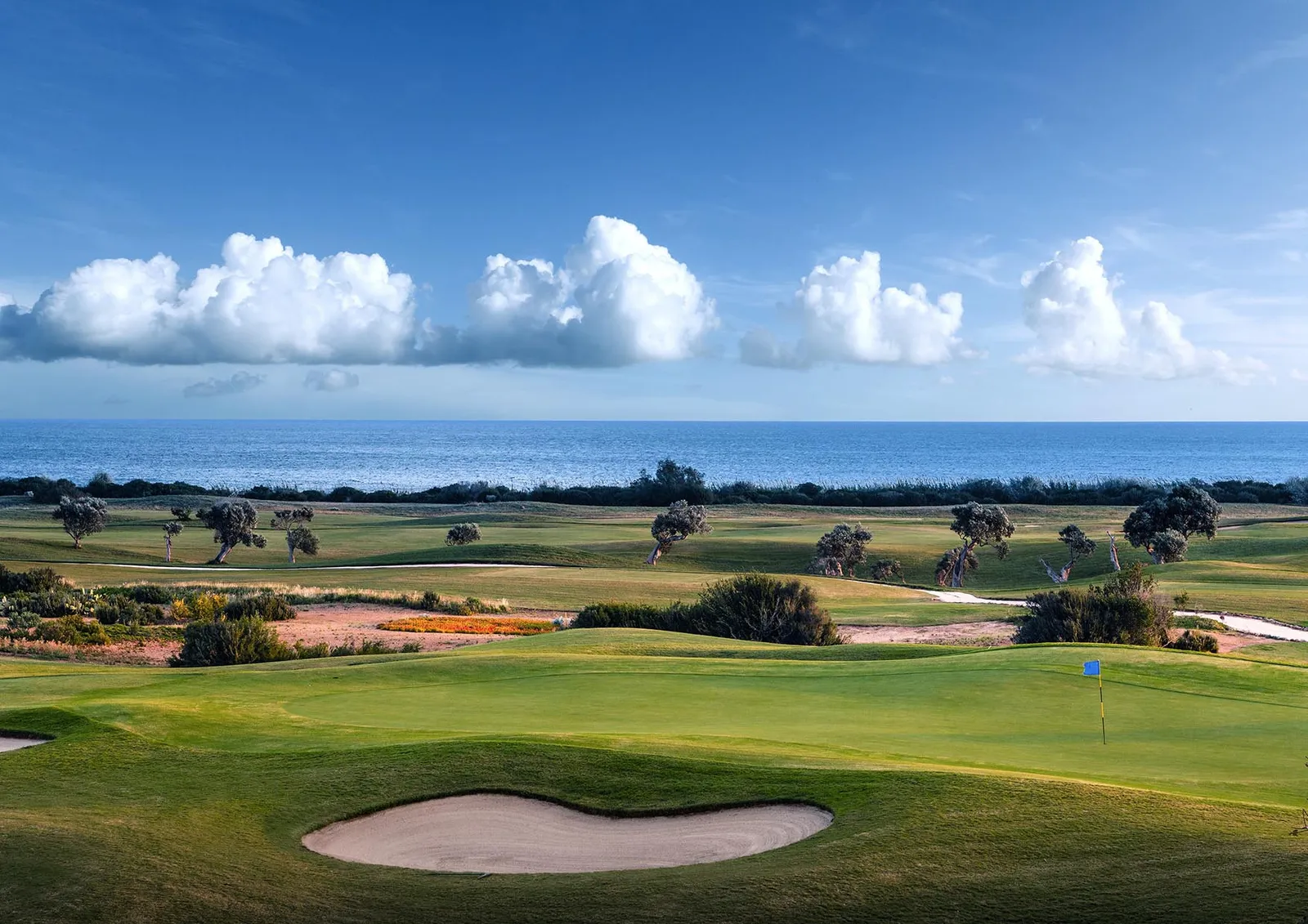 San Domenico Golf: a green with a bunker and a flag, fairways and scattered trees running down to the sea, a line of clouds over the horizon.