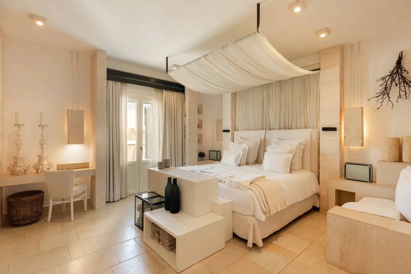 The La Egnazia suite: bed under a draped canopy, a writing desk with candlesticks, stone niches and a glazed door.