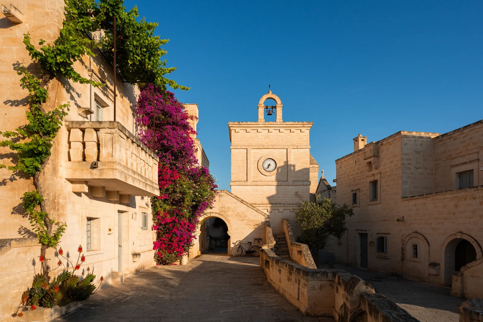 A lane in Il Borgo in daylight: a stone house with a balcony and bougainvillea on the left, and at the end the clock tower with its bell above an archway, a bicycle leaning beside it.