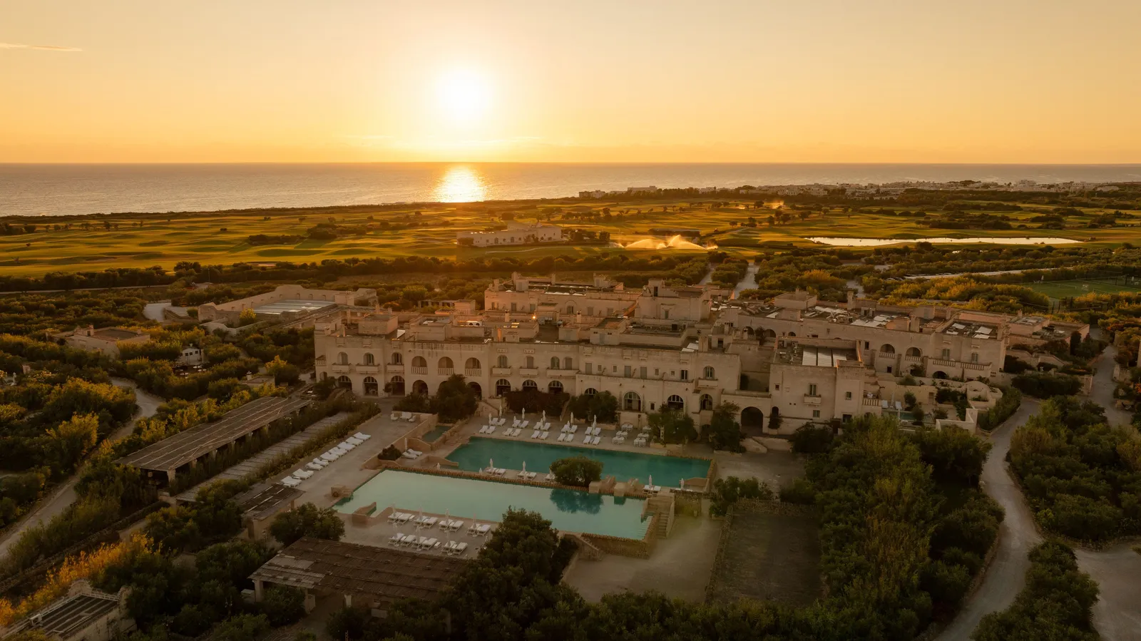 The estate from the air with the sun low over the sea: La Corte and its 2 pools in the foreground, the golf course and the coast beyond.