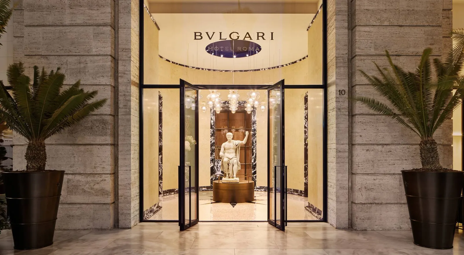The entrance at 10 Piazza Augusto Imperatore: glass doors open onto the round vestibule, with a seated statue of Augustus beneath glass pendant lamps; above, the lettering “Bvlgari Hotel Roma”, and a potted palm either side of the travertine doorway