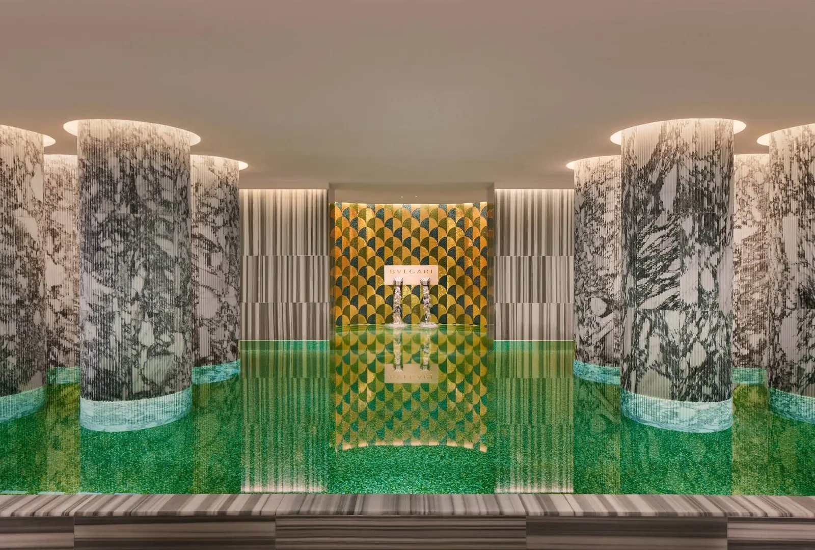 The spa pool: water over green mosaic, columns of white marble with dark veining and, at the far end, a gold and green mosaic wall with the Bvlgari name and 2 spouts of water