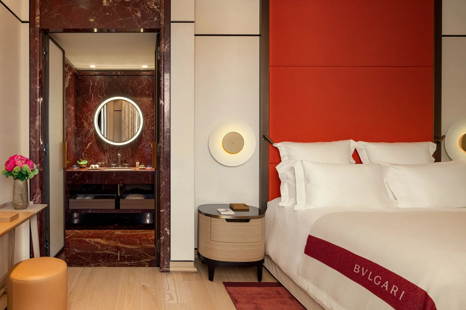 A Deluxe Room: a bed under a red headboard panel, a round lit mirror and a red-marble bathroom beyond