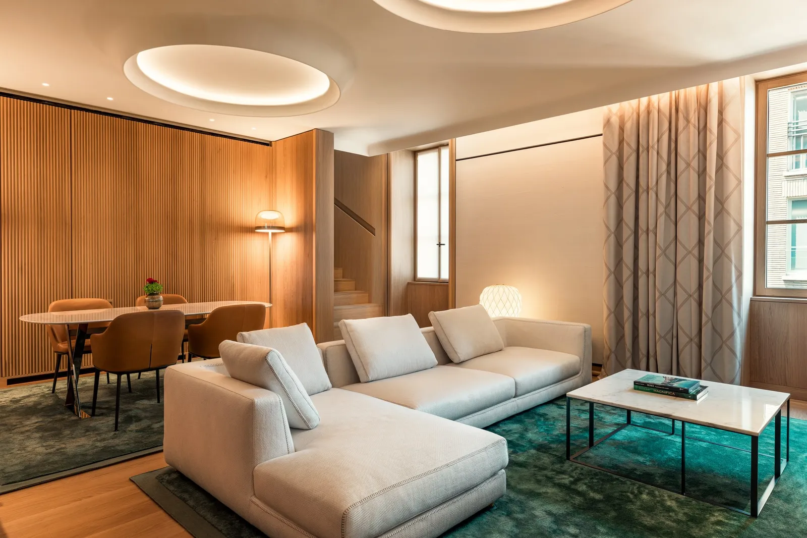 A Premium Suite living room: a white corner sofa on a green rug, a dining table and tall curtained windows