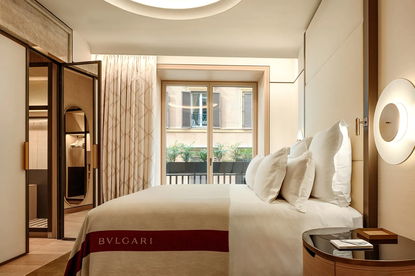 A Superior Suite bedroom: a bed with a red Bvlgari throw, a round wall lamp and a tall window onto the rooftops