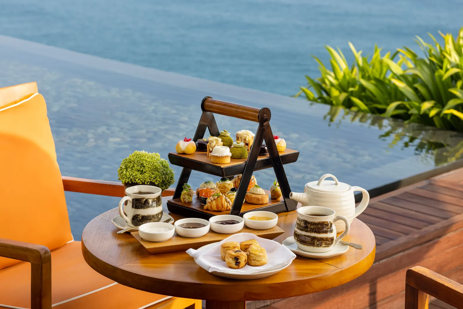 Afternoon tea at the edge of a pool above the ocean: a 3-tier stand of sweets and savouries, a teapot and scones.
