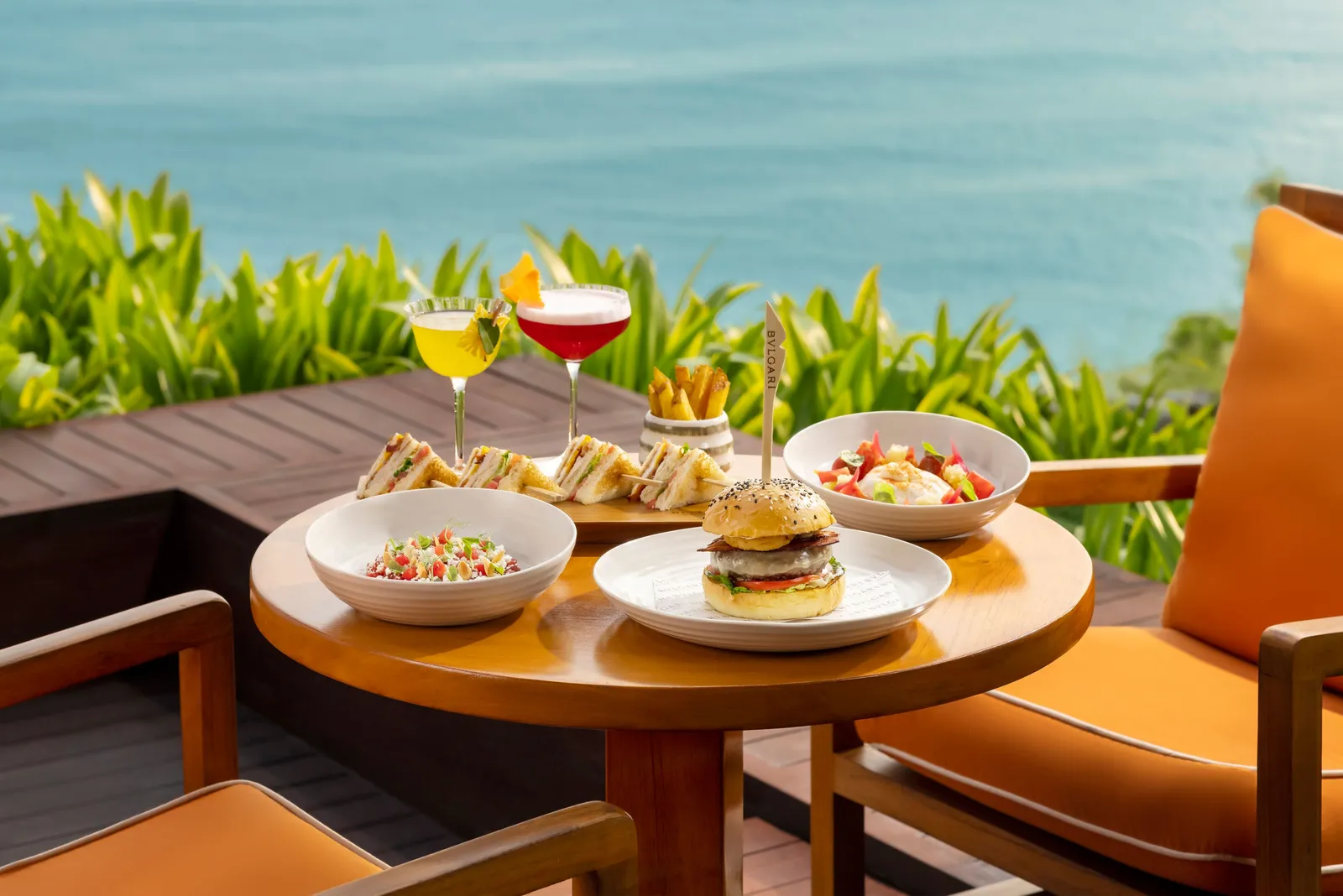 A table at the Bvlgari Bar above the ocean: 2 cocktails, sandwiches, a burger and salads, orange armchairs.