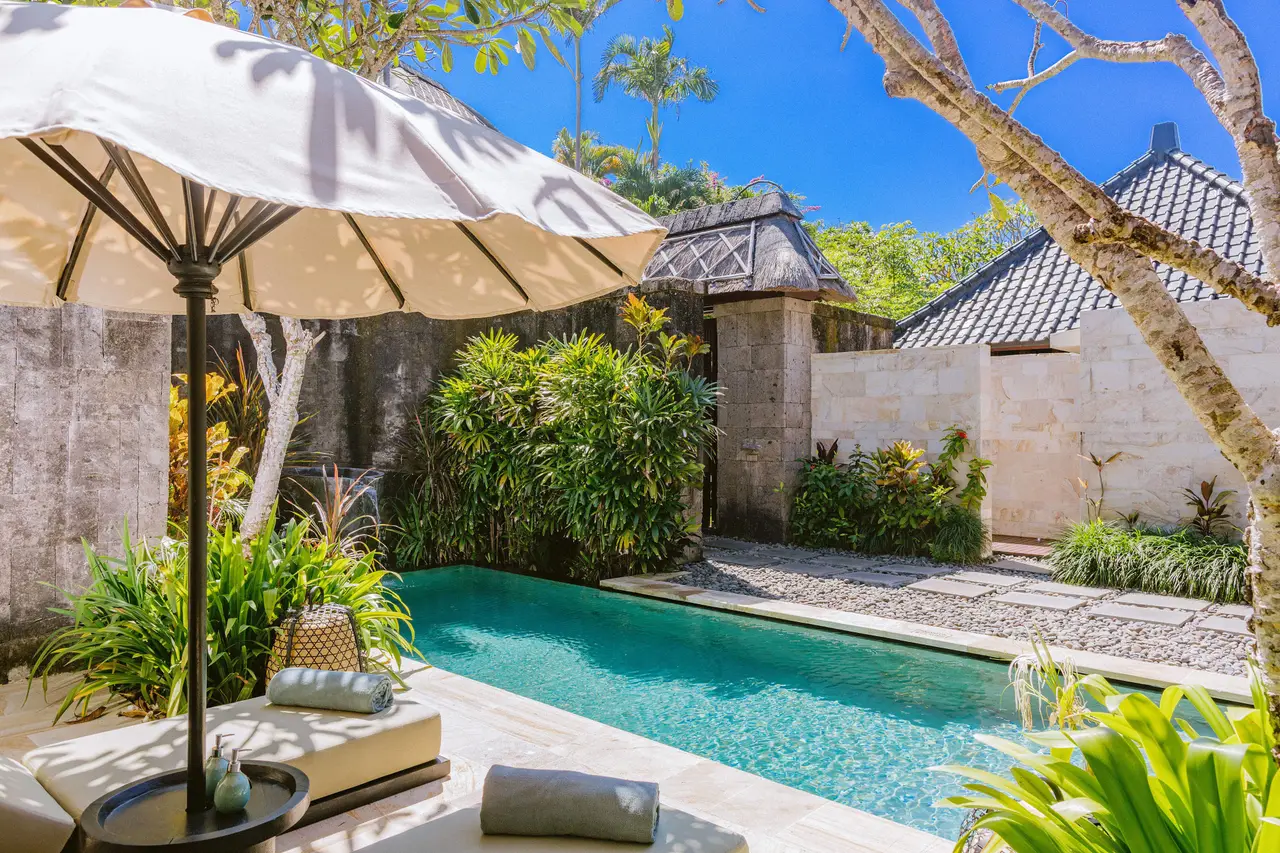 A villa’s private pool between stone walls and tropical plants: a lounger, a parasol and the roofs of other villas.