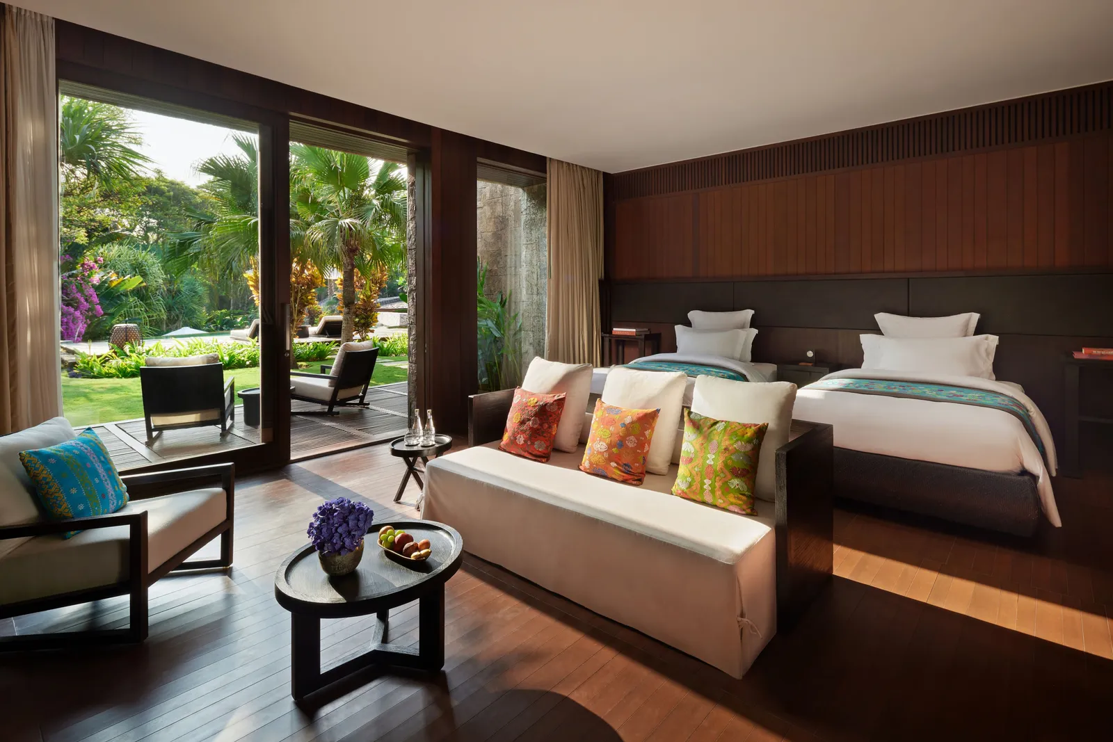 A Mansion bedroom with 2 beds, a sofa with bright cushions and glass walls onto the garden.