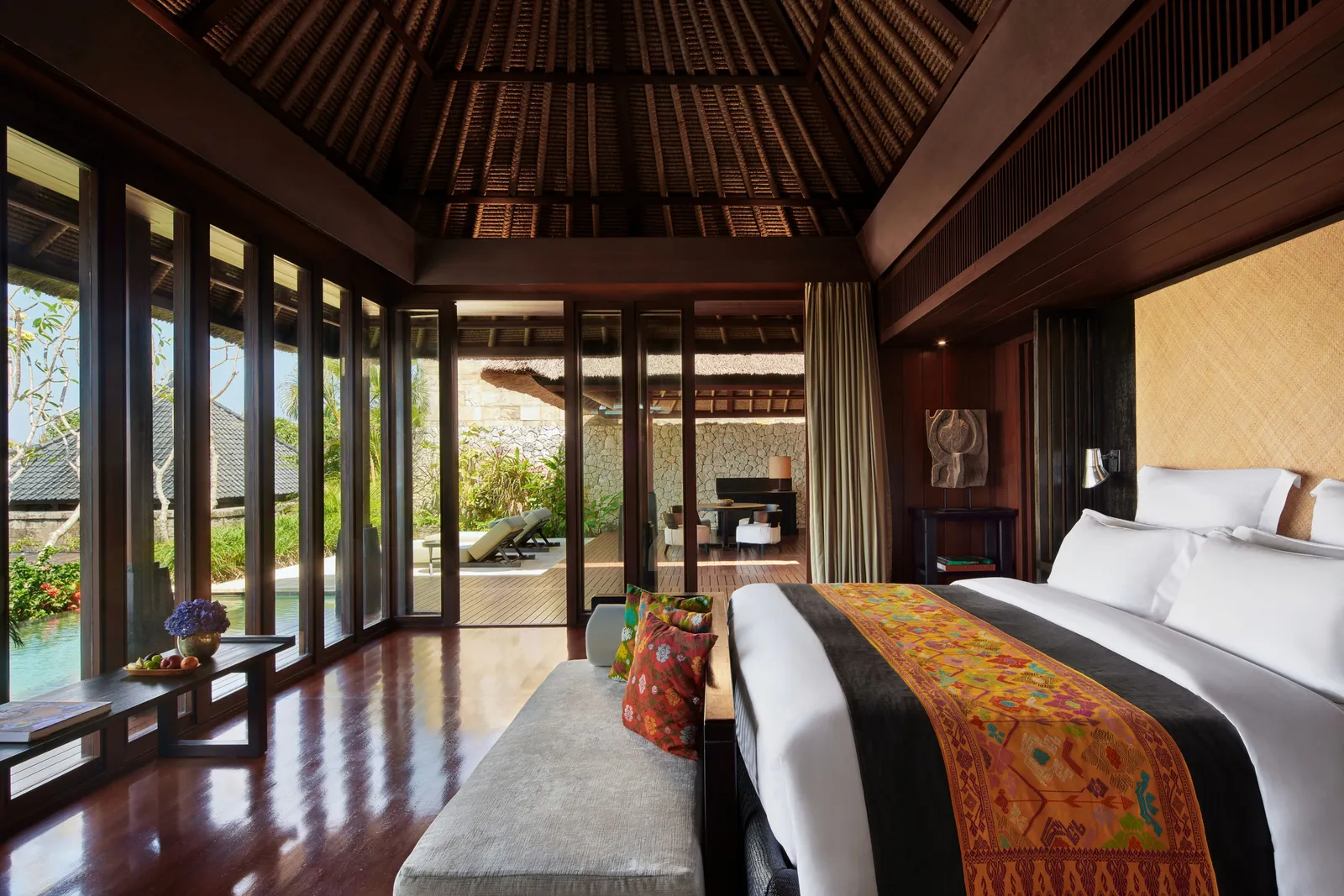 A villa bedroom: the bed with its songket runner, the pitched timber ceiling and the glass doors onto the pool.