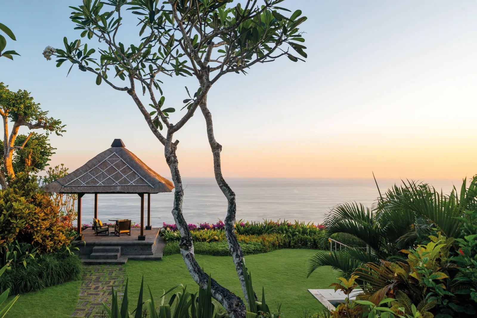 The garden of the Bvlgari Villa at sunset: a thatched pavilion on the lawn, 2 frangipani trees and the sea beyond.