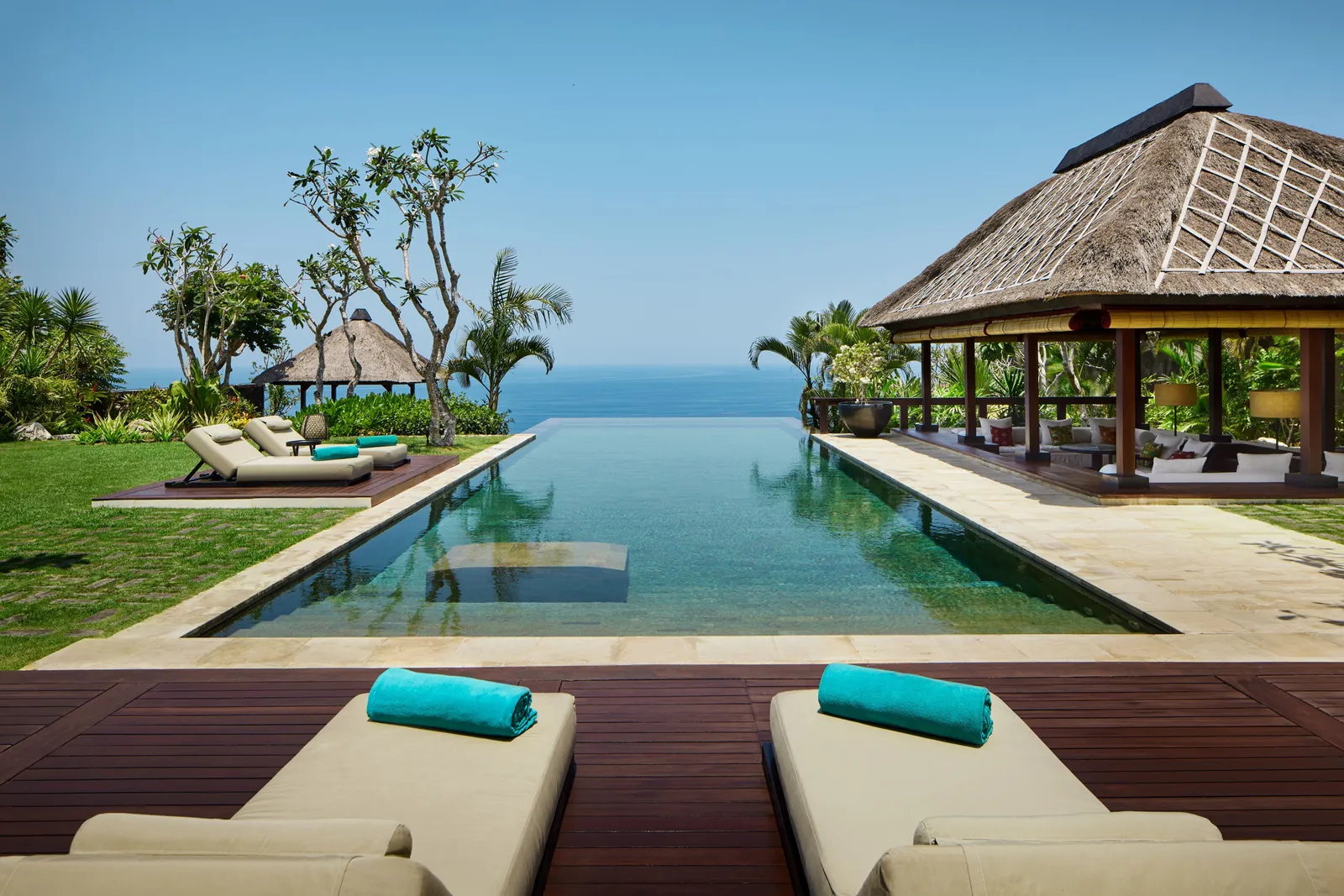 The Bvlgari Villa’s pool vanishing into the sky between 2 thatched pavilions, with 2 loungers in the foreground.