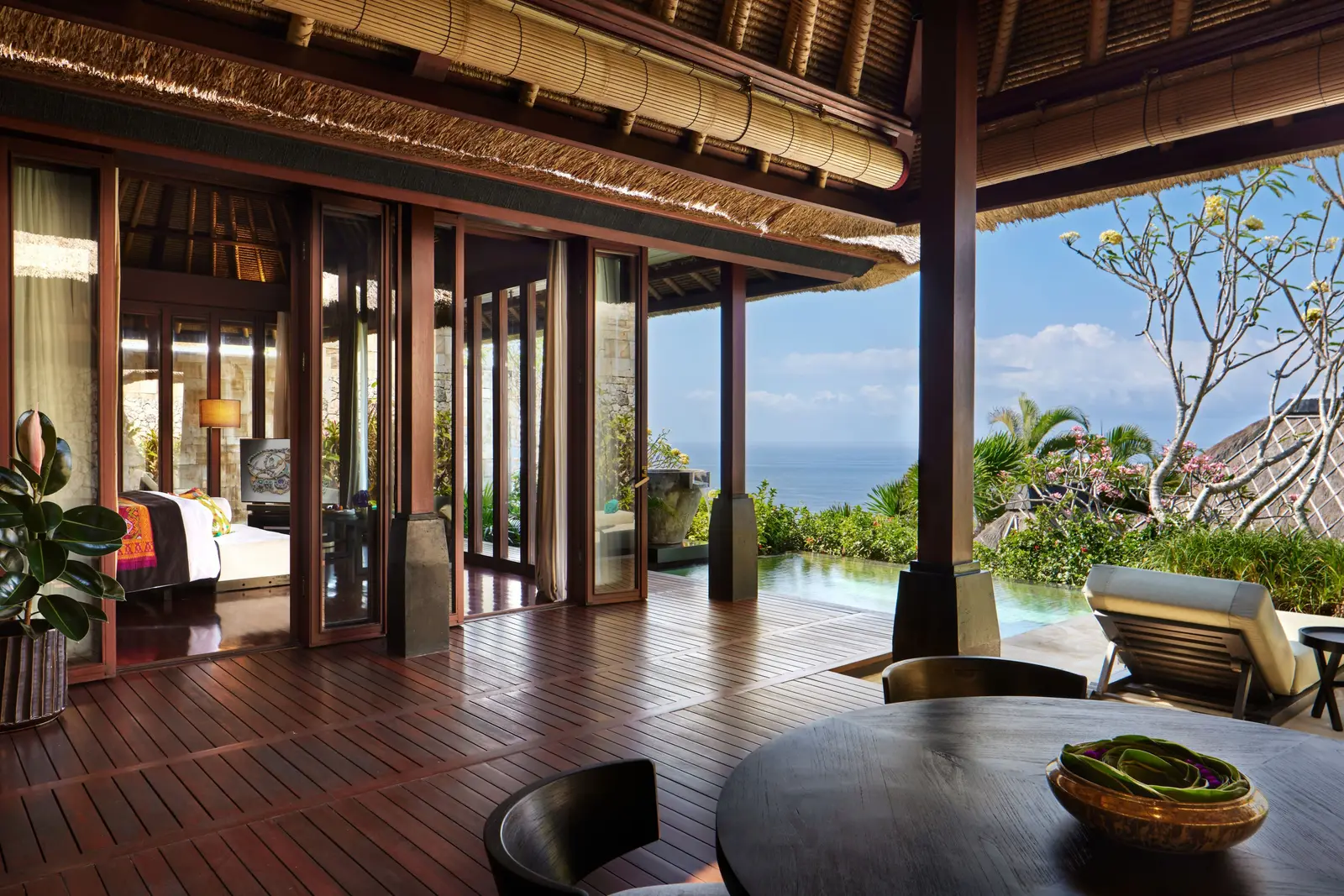 An Ocean View Villa’s veranda: the round wooden table, the loungers, the bedroom behind glass doors and the sea between frangipani.