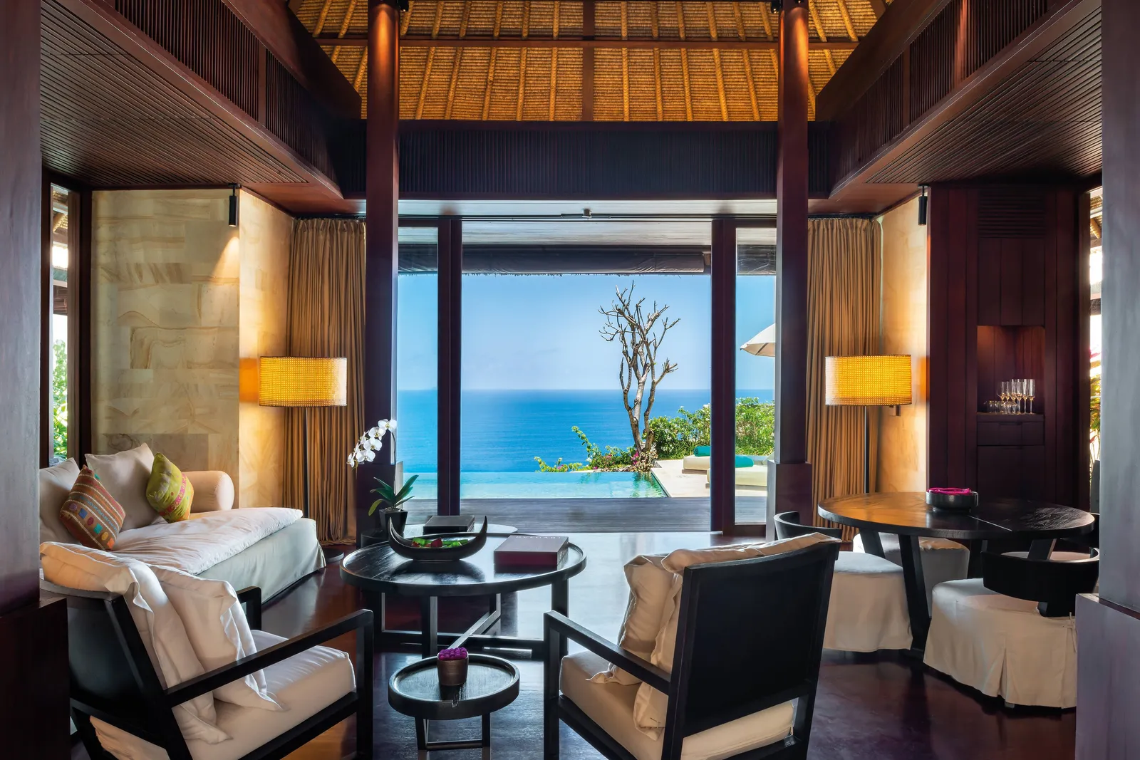 The living room of a 2-bedroom Ocean Cliff Villa: armchairs and a sofa under the timber roof, the door open on the pool and the sea.