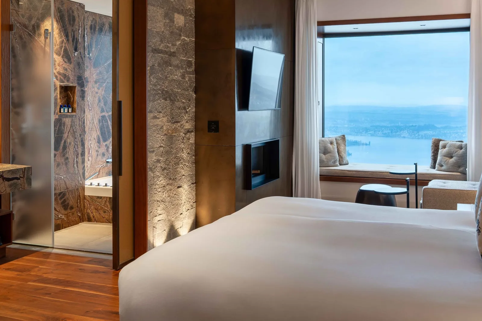 A Contemporary Lake View Bay Suite: the bed, a fireplace in the wall, a marble bathroom behind glass and the lake in the bay window