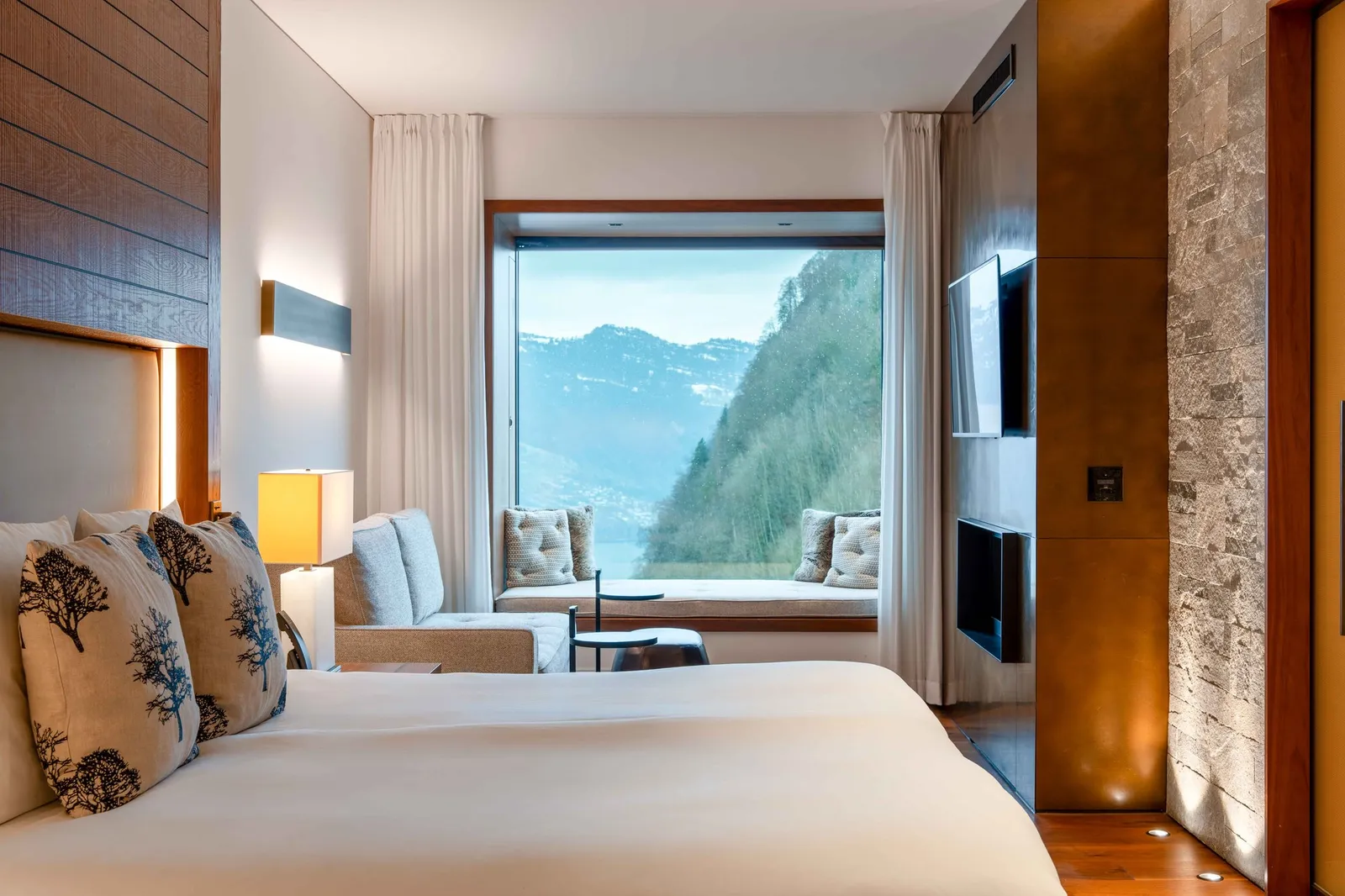 A Contemporary Deluxe Room: the bed, a window seat with cushions in the bay window over the lake and the cliff, and walls of stone and bronze